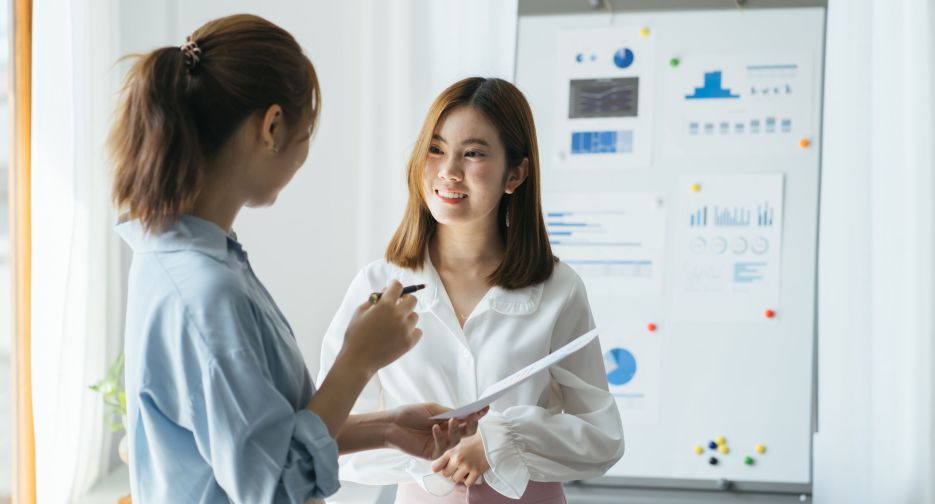 Two young businesswomen doing a presentation in a meeting or conference standing in front of a flip chart with hand drawn chart pointing to it with a pen